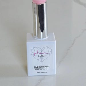 May include: A white rectangular bottle of nail polish with a silver cap and a pink brush. The bottle has the text "the glam LAB" and "RUBBER BASE" printed on it. The bottle contains 0.5 fl oz of product.