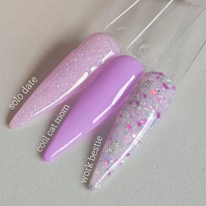May include: Three nail polish swatches on clear sticks. The first is a light pink with glitter, labeled "solo date". The second is a solid lavender, labeled "cool cat mom". The third is white with glitter and pink and silver flakes, labeled "work bestie".