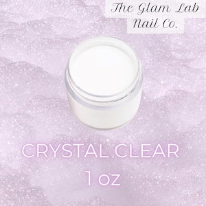 May include: A clear acrylic powder in a small, transparent jar. The jar is filled with white powder and sits on a light purple, glittery surface. Text on the image reads "Crystal Clear" and "1 oz.", with the brand name "The Glam Lab Nail Co."