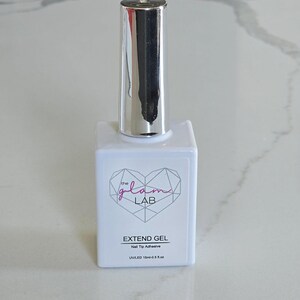 May include: A white bottle of nail tip adhesive with a silver cap. The bottle has a logo with the words "the glam LAB" inside a pink heart-shaped geometric design. The text "EXTEND GEL" and "Nail Tip Adhesive" are printed below the logo.