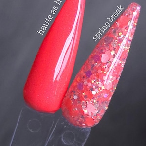 May include: Two nail polish swatches on clear sticks. One is a solid, bright red with the text "haute as hell-o". The other is a red glitter polish with the text "spring break". Both are against a dark grey background.