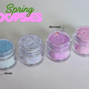May include: Four small, clear jars filled with colorful glitter. The jars contain blue, iridescent, purple, and pink glitter. Text above reads "Spring OOPSIES". The first jar is labeled "sun" and "shade", and the third jar is labeled "thermal".