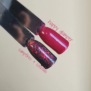 May include: Two nail polish swatches on black sticks. One is a deep red, labeled "happy glamper." The other is a dark base with pink and orange glitter, labeled "campfires & cocktails."