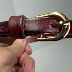 May include: A dark burgundy leather belt featuring a polished gold-tone metal buckle. The belt strap has a smooth, slightly distressed finish with visible stitching along the edges and a matching leather keeper loop near the buckle hardware.