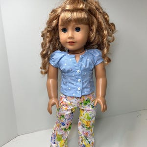 May include: A doll with long curly light brown hair and blue eyes. The doll wears a light blue short-sleeved shirt with white polka dots and three small white buttons down the front. The pants feature a colorful abstract paint splatter pattern.