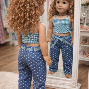 May include: A doll wearing a blue and green plaid crop top and blue jeans with white heart patterns. The doll is reflected in a white framed mirror. The doll has curly blonde hair.