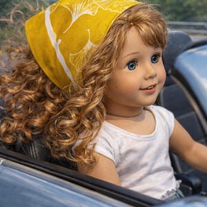 May include: A doll with blue eyes and curly red hair wearing a yellow headscarf with a white leaf pattern and a white t-shirt. The doll is seated in a blue convertible car.