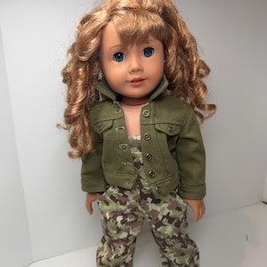 May include: A doll with long curly auburn hair and blue eyes wearing an olive green denim-style jacket with metal buttons over a pair of camouflage print pants. The doll has a neutral expression and is positioned in a standing posture.