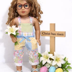 May include: A doll wearing patchwork overalls, holding white flowers, and wearing round glasses. A wooden cross with the text "Christ has risen" stands nearby, along with Easter eggs and lilies. The doll's outfit features pastel colors.