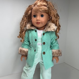 May include: A doll with long wavy brown hair and blue eyes wearing a mint green corduroy overall set and a matching mint green jacket with a faux fur collar and cuffs. The jacket features four brown buttons and the text JOE'S on the chest.