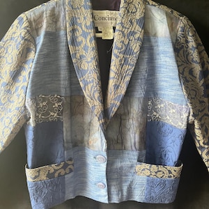 May include: A patchwork blazer featuring various shades of blue and tan with a floral jacquard pattern. The garment has a shawl collar, long sleeves, two front pockets, and two circular buttons. A label inside reads Concinne.