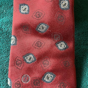 May include: A red tie with a repeating pattern of small, ornate shapes in navy, gold, and brown. The tie has a subtle sheen and a small, circular logo near the bottom.
