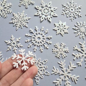 May include: A collection of white glitter snowflake embellishments of various sizes and designs. The snowflakes are scattered on a light blue background.