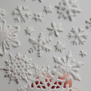 May include: A collection of white glitter snowflakes of various sizes. The snowflakes are scattered on a white background. One large snowflake is being held in a hand.