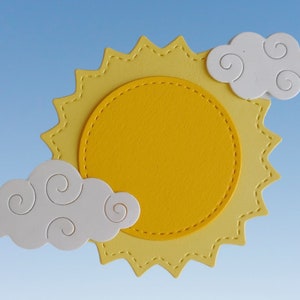 May include: A yellow sun with a white cloud on each side. The sun has a scalloped edge and a stitched circle in the center. The clouds have a swirl design.