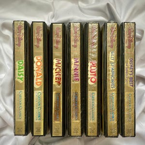 Peut inclure: Une collection de sept cassettes VHS Disney Cartoon Classics, avec des personnages tels que Daisy, Donald, Mickey, Minnie, Pluto, Silly Symphonies et Disney's Best. Les cassettes ont un emballage doré et noir avec le logo Disney.