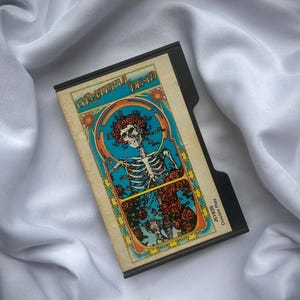 May include: A vintage cassette tape featuring a colourful image of a skeleton with flowers and the text "Grateful Dead" on the front. The tape is labelled "1973" and "Don't Play Dead".