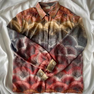 May include: A patterned button-down shirt in shades of red, orange, brown, grey, and white. The shirt has a gradient design with a repeating tree-like pattern. It features a pointed collar and buttoned cuffs on the long sleeves. The fabric appears lightweight.