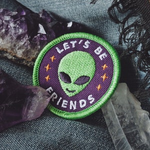 Op de afbeelding: Groen alien geborduurd patch met de tekst "Let's Be Friends" in wit op een paarse achtergrond. De patch is omgeven door een groene rand en heeft drie oranje sterren.