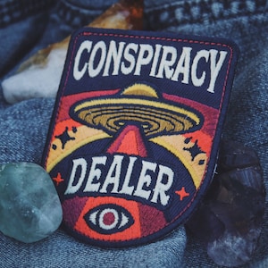 Op de afbeelding: Een geborduurde patch in blauw, rood en geel met een UFO en de tekst "Conspiracy Dealer" en een enkel oog met een rode pupil.