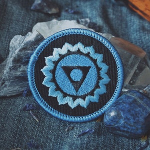 May include: A blue and black embroidered patch with a stylized chakra symbol. The symbol is a triangle with a circle inside, surrounded by a flower-like design. The patch is on a denim background with blue and white stones.