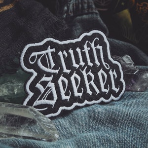 May include: Black and white embroidered patch with the words "Truth Seeker" in a gothic font.