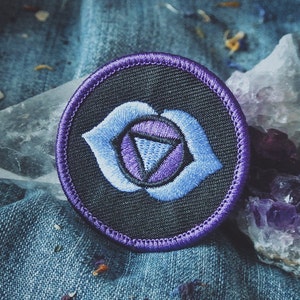 May include: A purple and blue embroidered patch with a third eye symbol, representing the sixth chakra, the Ajna chakra. The patch is on a denim background with crystals.