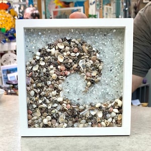 May include: A white framed square wall art piece with a wave design made of seashells and small pieces of glass.