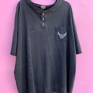 Puede incluir: Una camisa negra de manga corta con botones y un bolsillo en el lado izquierdo. El bolsillo tiene un logotipo de Harley Davidson con un diseño de águila. La camisa tiene un cuello abotonado.