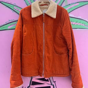 May include: An orange corduroy jacket with a cream-colored sherpa collar and cuffs. The jacket has a zip-up front, two front pockets, and is hanging on a wooden hanger. The background is a pink wall with a palm tree design.