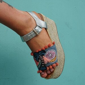 May include: Silver wedge sandals with a blue and white embroidered design featuring a palm tree, watermelon, and citrus fruit. The sandals have a gold platform and a buckle closure.