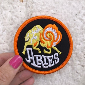 Op de afbeelding: Een geborduurde zwarte en oranje patch met een gele ram en het woord "Aries" in wit.