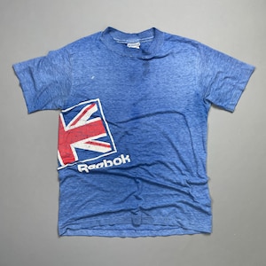 May include: A blue t-shirt with a faded, distressed look. The shirt features a white Reebok logo and a red, white, and blue Union Jack flag graphic.