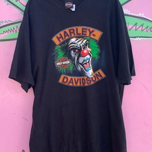 May include: Black t-shirt featuring a graphic of a clown wearing a Harley Davidson patch. The clown has green hair, a red nose, and a wide grin. The text "Harley Davidson" is printed in orange and black.