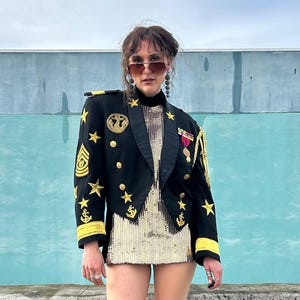May include: A woman wearing a black military-style jacket with gold stars and anchors, a gold sequined mini dress, and sunglasses.