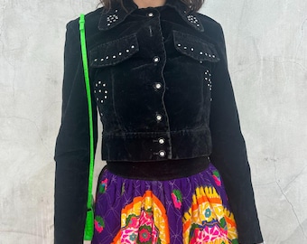 May include: Black velvet jacket with white stud accents and a button-up front. The jacket is paired with a vibrant, patterned skirt in purple, orange, and green. The outfit is accessorized with rhinestone sunglasses and a neon green bag.