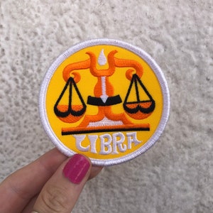Op de afbeelding: Een geborduurde patch in geel en wit met het sterrenbeeld Weegschaal en het woord "LIBRA" in witte letters.