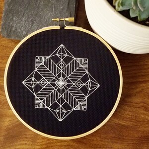 May include: A geometric embroidery hoop art piece. The design features a white, symmetrical pattern on a black fabric background. The embroidery is framed by a natural wood hoop with a brass clasp. The artwork is displayed on a wooden surface.