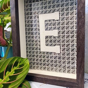May include: A framed cross-stitch piece with a black and white pattern of stylized pine trees. The letter "E" is stitched in white against the background pattern.