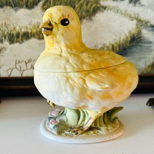 May include: A ceramic chick figurine with a lid, painted in shades of yellow and cream. The chick is perched on a white base with green leaves and pink flowers. The figurine has black eyes and a detailed beak.