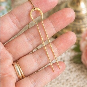 May include: A gold-toned metal hair fork featuring a U-shaped design with a textured, leaf-like pattern at the top and two slender, pointed prongs. A delicate gold chain is attached to the top centre of the hair fork, extending upwards.