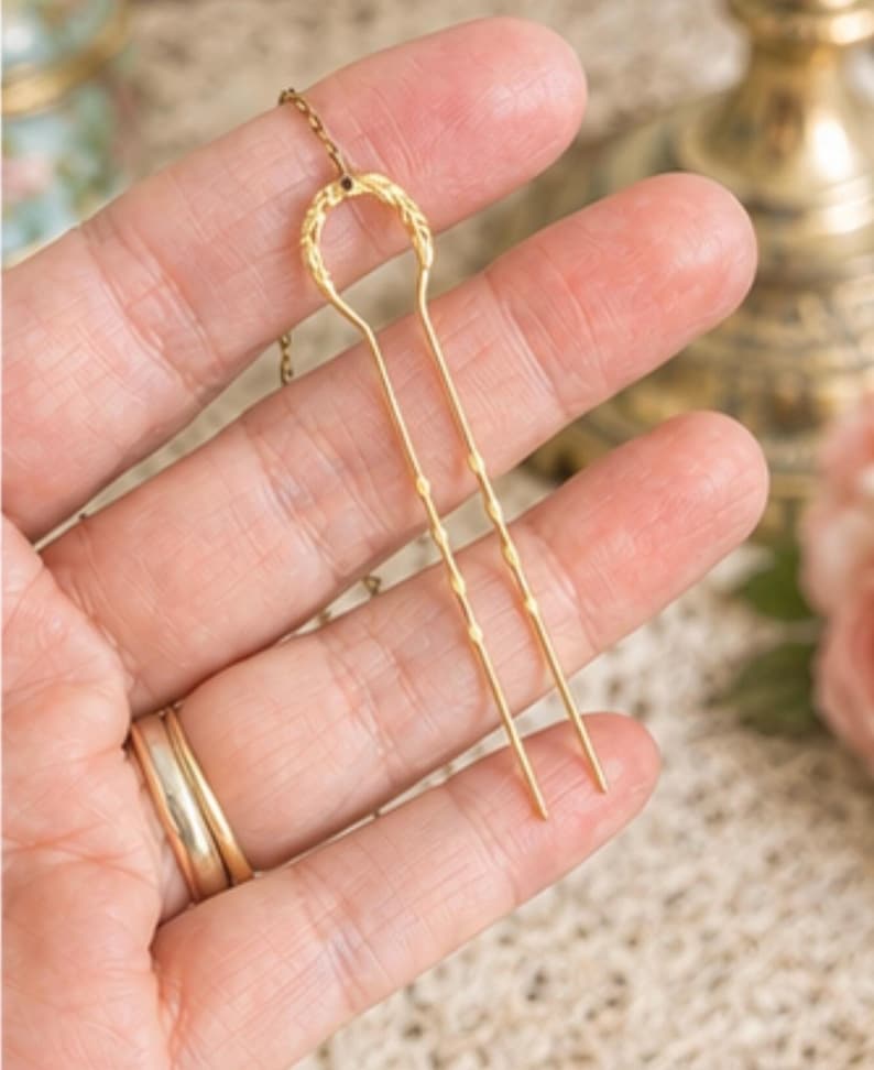 May include: A gold-toned metal hair fork featuring a U-shaped design with a textured, leaf-like pattern at the top and two slender, pointed prongs. A delicate gold chain is attached to the top centre of the hair fork, extending upwards.