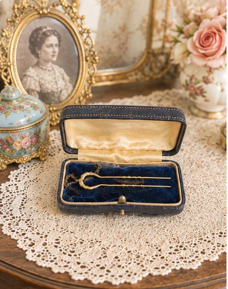 May include: A gold-toned metal hair fork with a U-shaped design and textured detailing, resting inside a rectangular navy blue hinged jewellery box. The interior of the box is lined with deep blue velvet and features a cream-coloured satin lid lining.