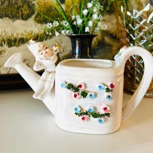 May include: A white ceramic watering can with a cherub figurine and floral accents. The watering can has a long spout, a curved handle, and is decorated with clusters of blue, pink, and green flowers. The cherub is perched on the side of the can.