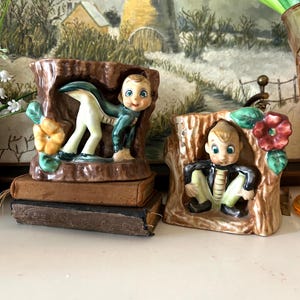 May include: Two vintage ceramic figurines. One shows a boy in a green jacket and white pants, emerging from a tree trunk. The other shows a boy in a black jacket and green pants, sitting in a tree trunk. Both have blue eyes and are on top of old books.