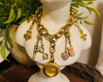 May include: A gold charm necklace featuring a watch, heart, cross, and other decorative charms. The necklace is displayed on a white bust, with foliage in the background. The chain has a chunky link design.