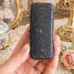 May include: A rectangular, dark navy blue jewellery box with a textured, pebbled exterior finish. The small, elongated case features visible metal hinges along the side and a worn, aged appearance with slight scuffing on the edges and surface.