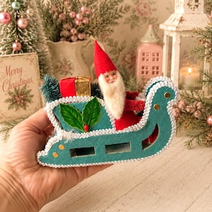 May include: A vintage-style Santa Claus figurine seated in a teal felt sleigh. The Santa has a white beard, a red hat, and red clothing. The sleigh is decorated with white trim, gold studs, and green holly leaves. A red and gold gift box sits behind.