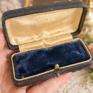 May include: A rectangular jewellery box with a dark textured exterior, a hinged lid, and a gold-toned metal latch. The interior features a cream-coloured satin lining on the lid and a deep blue velvet insert with a slot for a ring or small item.