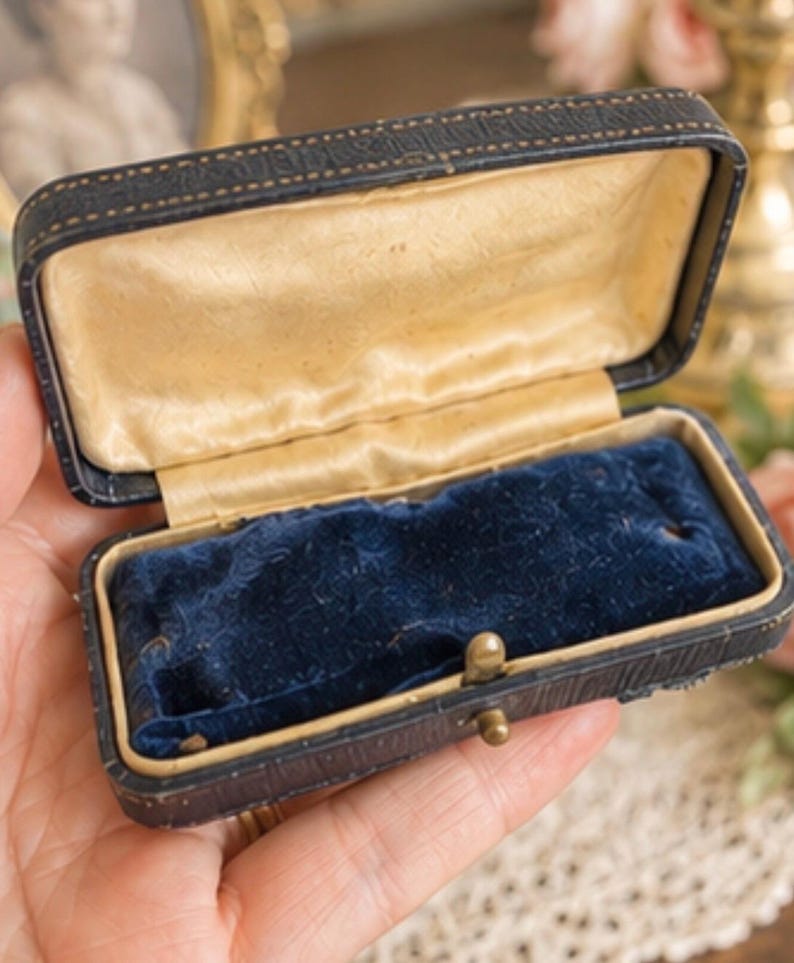 May include: A rectangular jewellery box with a dark textured exterior, a hinged lid, and a gold-toned metal latch. The interior features a cream-coloured satin lining on the lid and a deep blue velvet insert with a slot for a ring or small item.